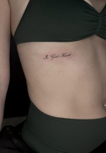 Elegant script tattoo reading "In Glori Honde" on side ribcage.