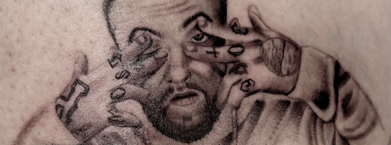 A black and grey portrait of a man with tattoos on his hands, making a face.